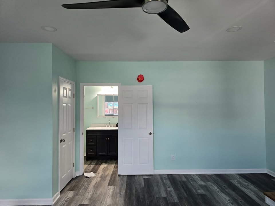Room with dark gray plank flooring, light green walls, white trim and a ceiling fan, opening to a bathroom vanity